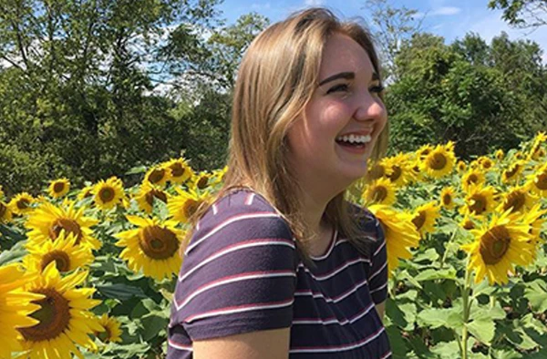 Smiling woman in a striped shirt standing in a field of sunflowers under a clear blue sky.